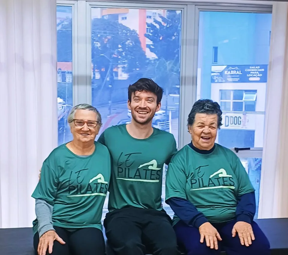 Dona Maria e Dona Isabel, alunas do LE Pilates Studio, sorrindo ao lado de um professor, todos de camiseta verde do estúdio